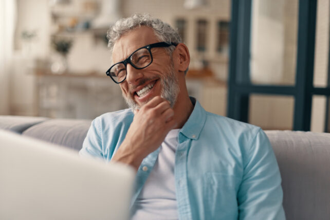 Homem alegre sênior em roupas casuais e óculos, usando laptop e sorrindo enquanto está sentado no sofá