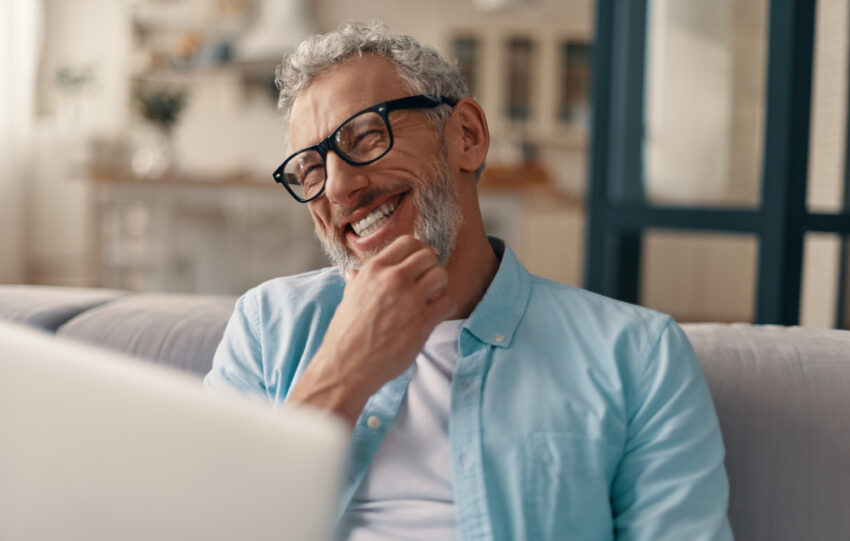 Homem alegre sênior em roupas casuais e óculos, usando laptop e sorrindo enquanto está sentado no sofá